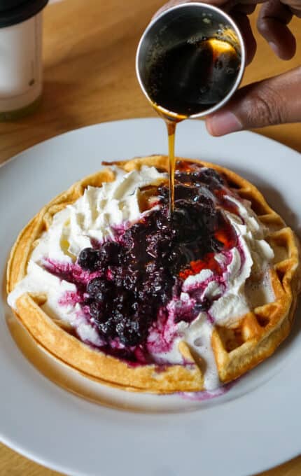 A waffle on a white plate topped with whipped cream and berry compote. A hand is pouring syrup from a small cup onto the waffle. A coffee cup is partially visible in the background on a wooden table.