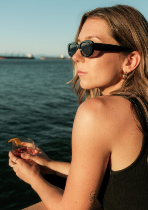 A person wearing sunglasses and a black tank top holds a drink while looking over a calm body of water with ships in the background under a clear sky.