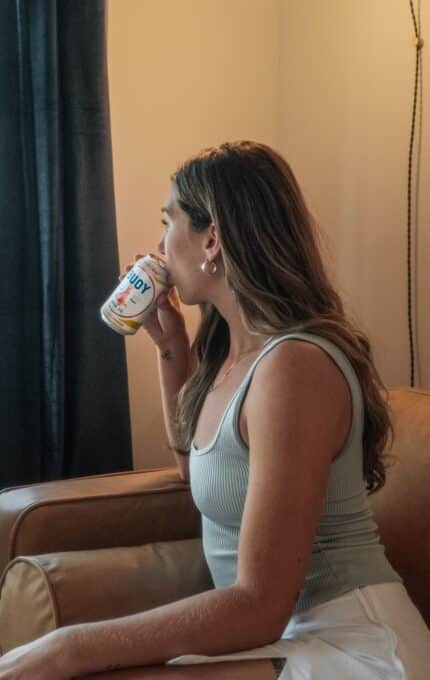 A person with long hair sits on a tan leather chair, looking out a window while drinking from a can. The room has cream walls and dark curtains, and a hanging light is visible.