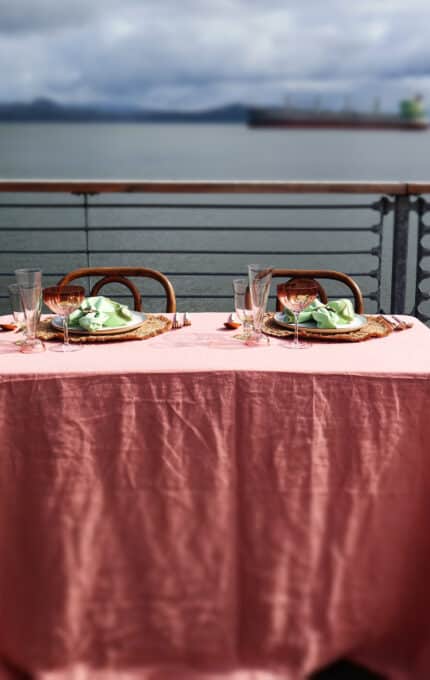 A table with a pink tablecloth is set for two with plates, napkins, and glasses. It is located outdoors on a deck overlooking a large body of water with ships in the distance. The sky is cloudy.