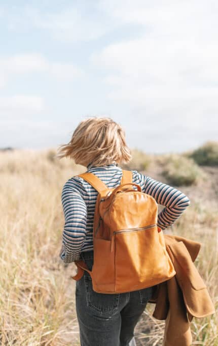 A person with short blonde hair, wearing a striped shirt and jeans, carries a brown backpack while walking through a grassy field. They are holding a brown jacket and heading toward an overcast sky in the distance.