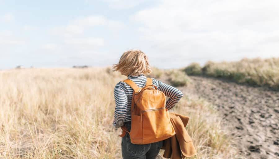 A person with short blonde hair, wearing a striped shirt and jeans, carries a brown backpack while walking through a grassy field. They are holding a brown jacket and heading toward an overcast sky in the distance.