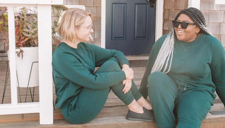 Two people sit on a wooden porch, both wearing matching green outfits. One is sitting cross-legged on the porch floor, and the other is sitting on the step, smiling. There is a potted plant and a blue door in the background.