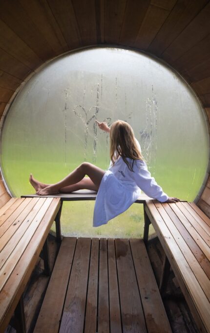 A person in a white robe sits inside a wooden sauna with a circular window. They are drawing on the fogged-up glass. The interior features wooden benches and a wooden floor. Outside the window, greenery is visible.