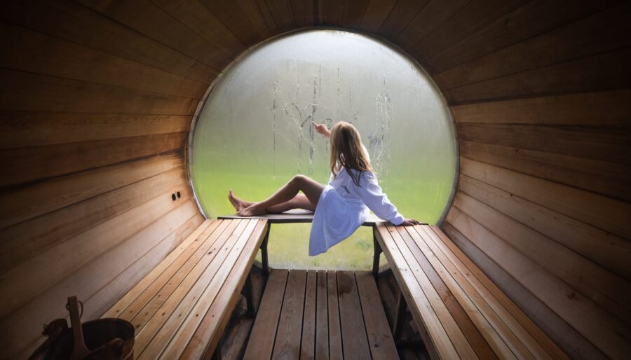 A person in a white robe sits inside a wooden sauna with a circular window. They are drawing on the fogged-up glass. The interior features wooden benches and a wooden floor. Outside the window, greenery is visible.