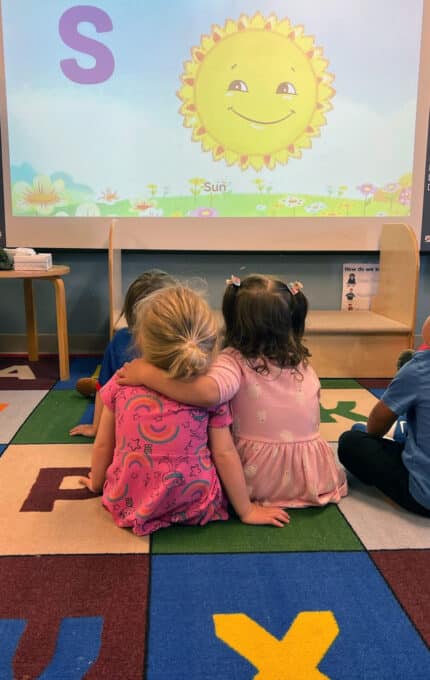 Four young children sit on a colorful alphabet mat, facing a screen showing a smiling sun and the letter "S". Two girls in pink sit close together, with one wrapping her arm around the other. The setting appears to be a classroom.