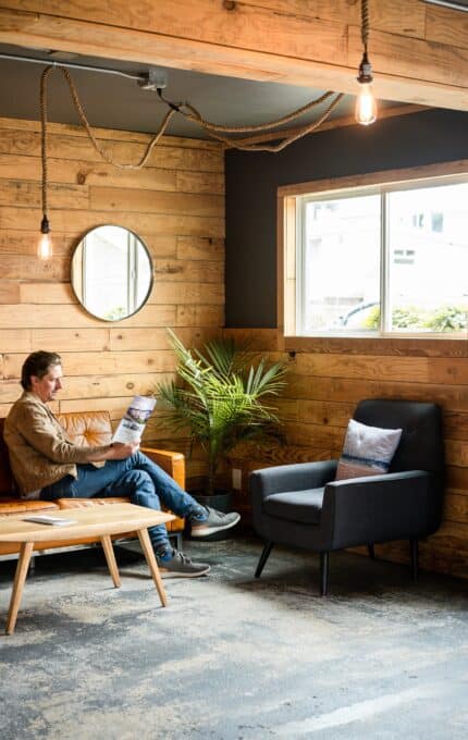 A person is sitting on a brown sofa reading a newspaper in a cozy room with wood-paneled walls. The room features a large window, a small round mirror, a gray armchair, a palm plant, and hanging light bulbs.