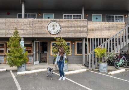 A woman with long hair, wearing a denim jacket and jeans, stands outside a building labeled "Check in and chill out" holding a drink. She is with a black and white dog. Bicycles and plants are nearby, and an outdoor staircase leads to the upper floor.