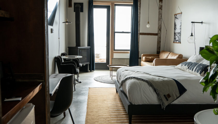 A modern bedroom with a large bed, a brown leather sofa, and a flat-screen TV. The room features concrete floors, a striped rug, and high ceilings with exposed beams. Natural light enters through a large window near the sofa.