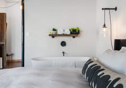 A bedroom with a white duvet-covered bed in the foreground. A black-and-white pillow sits on the bed. Behind the bed, there's a freestanding bathtub with a wooden shelf above it holding rolled towels and plants. A hanging light fixture is on the wall.