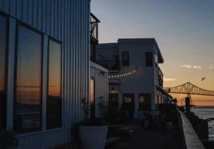 A waterfront building with large windows is visible at sunset. The reflections in the windows mirror the orange sky. String lights are strung along the building's facade. A bridge and a bird in flight are seen in the background.