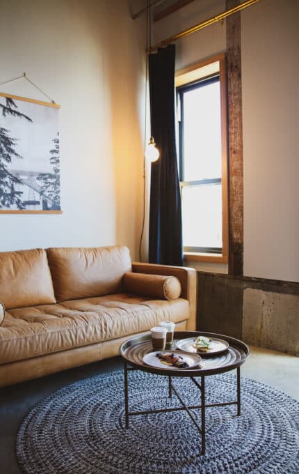 A cozy living room features a tan leather sofa, a round coffee table with plates and a cup, and a textured gray rug. A large window with black curtains lets in light, and a framed image hangs on the wall above the sofa.