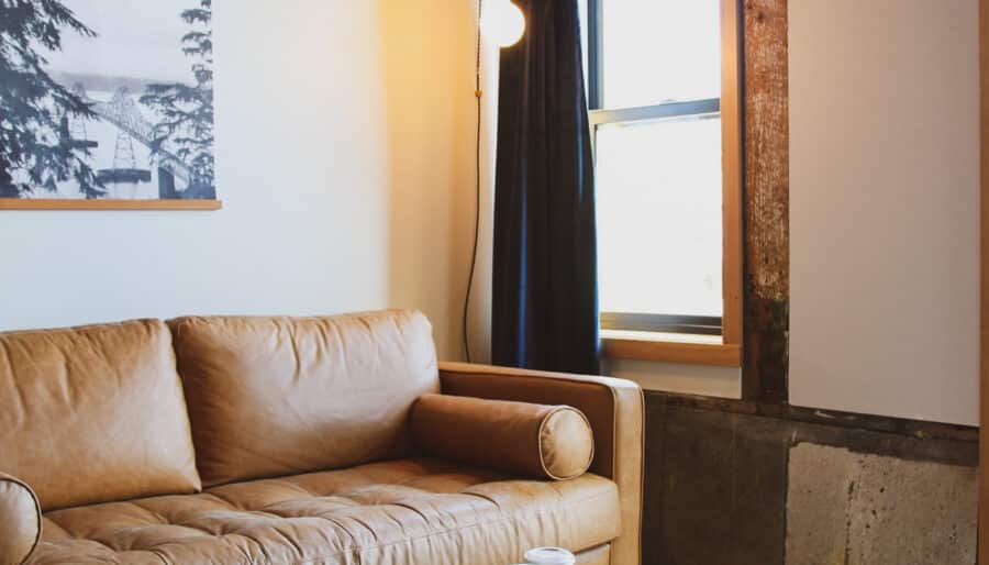 A cozy living room features a tan leather sofa, a round coffee table with plates and a cup, and a textured gray rug. A large window with black curtains lets in light, and a framed image hangs on the wall above the sofa.