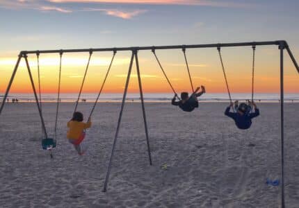 Three children swing on a swing set at the beach during sunset. The sky is streaked with shades of orange and pink, and the ocean is visible in the background. The sand is dotted with footprints.