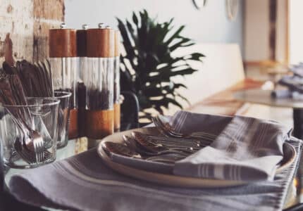 A restaurant table is set with neatly folded napkins and cutlery on a plate. A set of clear pepper mills is positioned behind the plate. A plant is visible in the background, adding a touch of greenery to the rustic decor.