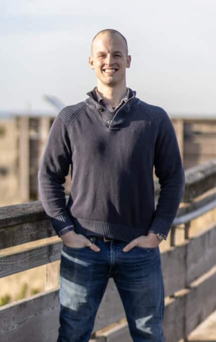 A man stands smiling on a wooden walkway with his hands in his pockets. He is wearing a dark sweater and jeans. The background shows a blurred natural landscape with a clear sky.