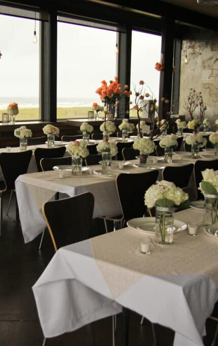 A spacious dining room is set up for an event, featuring long tables with white tablecloths and white floral centerpieces. Large windows offer a view of a beach, and hanging lights illuminate the space.