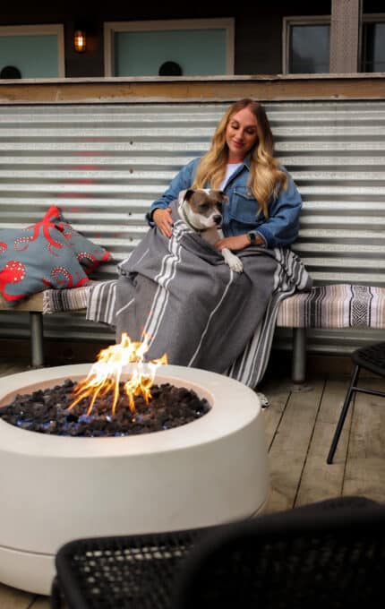 A woman sits on an outdoor bench, wrapped in a grey blanket with a bulldog on her lap. A circular fire pit with flames burns in front of her. A red and grey bag with crab motifs is placed beside her. She appears relaxed and content.