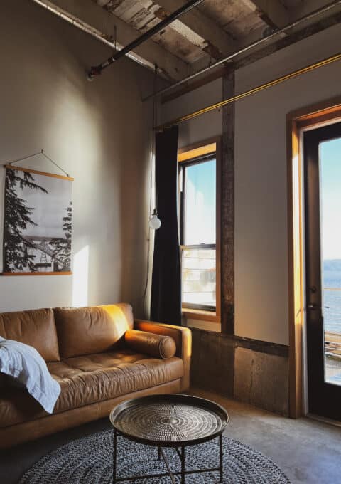 A cozy living space with a brown leather couch, a round metal coffee table on a textured rug, and large windows letting in natural light. A black-and-white wall hanging featuring a landscape is above the couch, with a view of water visible outside.