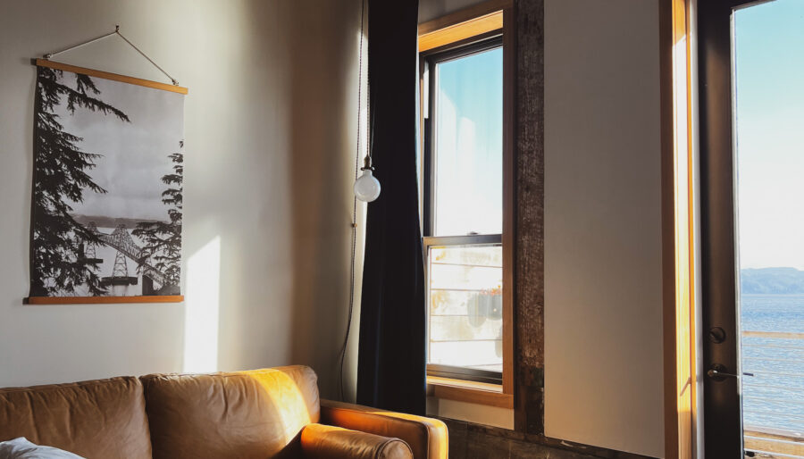 A cozy living space with a brown leather couch, a round metal coffee table on a textured rug, and large windows letting in natural light. A black-and-white wall hanging featuring a landscape is above the couch, with a view of water visible outside.