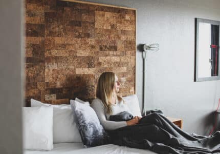 A woman with long blonde hair sits on a bed, wrapped in a dark blanket. She is gazing out a window. The room has a wooden accent wall, a grayish tone, and a floor lamp beside the bed. Two white pillows and one patterned pillow are visible.