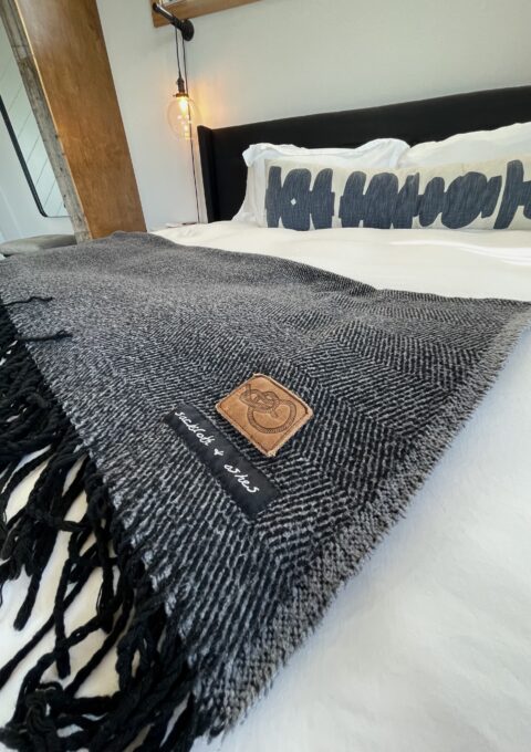 A bed with white sheets and a gray woven blanket featuring fringe and a leather patch is shown. The headboard is wooden, and there's a pillow with stylized text. A decorative light bulb hangs beside the bed.