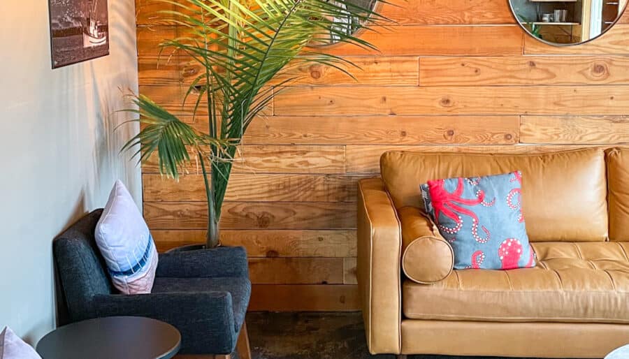 A cozy seating area with a tan leather sofa adorned with a decorative pillow, two dark upholstered chairs with light pillows, a small round table, a potted plant, and wooden walls featuring two round mirrors. A framed photo is hung on the wall.