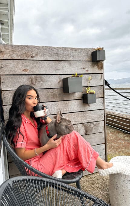 A person in a pink outfit sits on a patio chair, holding a small dog on their lap. They are drinking from a mug. The setting includes wooden panels with plants and a view of water and sky in the background.