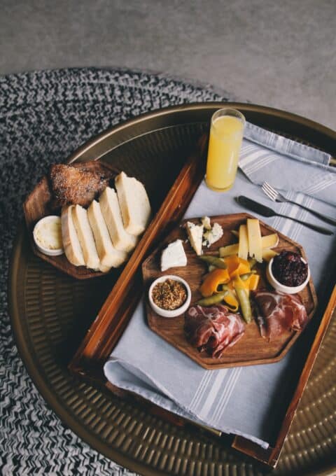 A tray on a round table contains a charcuterie board with various cheeses, cured meats, pickles, and spreads. Sliced bread and butter are on the side. A glass of orange juice is also on the tray, with a fork and knife placed nearby.