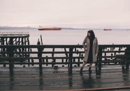 A person wearing a long coat stands on a wooden dock overlooking a body of water. Several ships are visible in the distance. The sky is overcast, and the dock appears wet, suggesting recent rain.