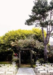 A garden setting with white chairs arranged in rows on either side of a gravel path leading to a floral archway. The arch is adorned with light pink flowers. Trees and greenery surround the area, creating a serene atmosphere.