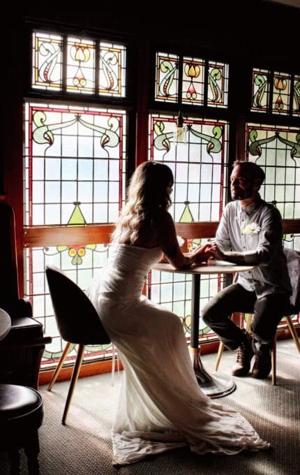 A bride and groom sit at a small round table by a large window with intricate stained glass. The bride is wearing a white dress and the groom is in a light-colored shirt and pants. They are holding hands and looking at each other.