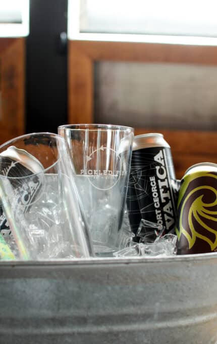 A metal bucket filled with ice holds two empty glass pint glasses and two beverage cans, one labeled "Vortex" with a distinctive design. The background features wooden and glass elements.