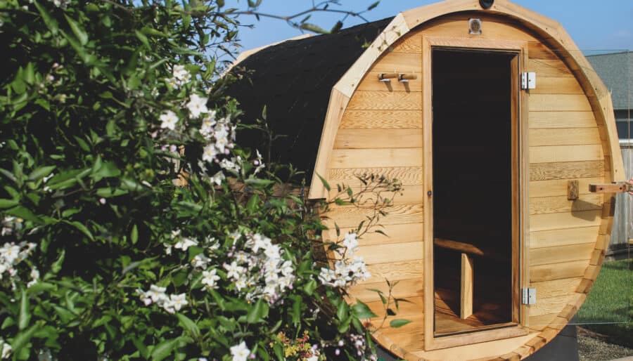 A wooden barrel-shaped sauna with an open door stands outdoors. Green foliage and white flowers are in the foreground, partially obscuring the view of the sauna. The sky is clear and blue.