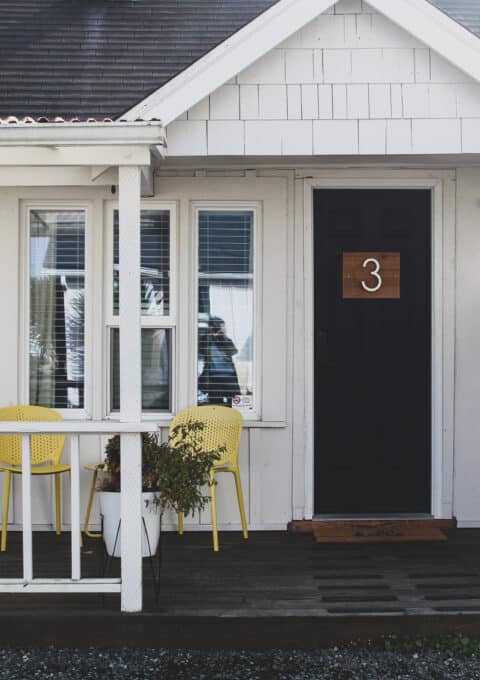 A small white house with a black door marked with the number 3. There are two yellow chairs and a small table on the front porch, along with a potted plant. The house has a pitched roof and gravel path leading to the entrance.