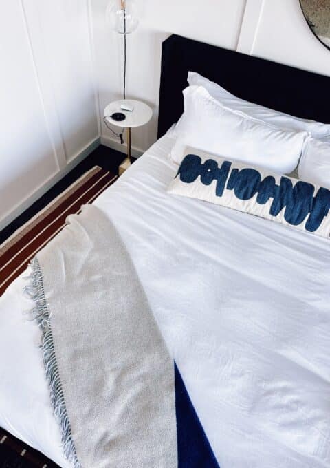 A neatly made bed with white sheets, two pillows, and a decorative blue and white pillow is shown. A beige throw blanket is draped over the bottom corner. A small round side table with a lamp and a clock is next to the bed.