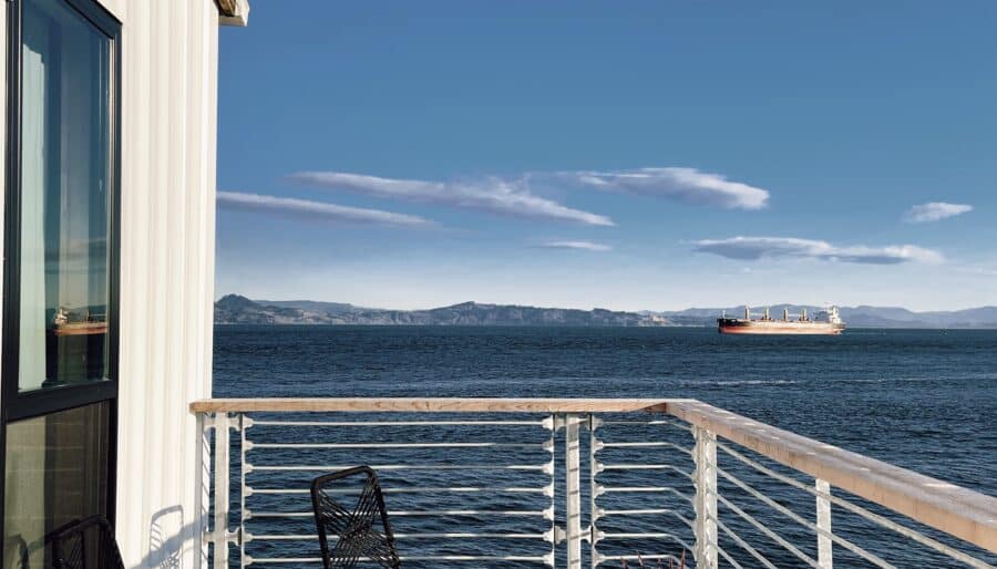 A sunny view from a wooden deck overlooking the ocean. The deck features two black metal chairs and a small table. A potted plant is positioned near the railing. A cargo ship is visible on the horizon under a clear blue sky.