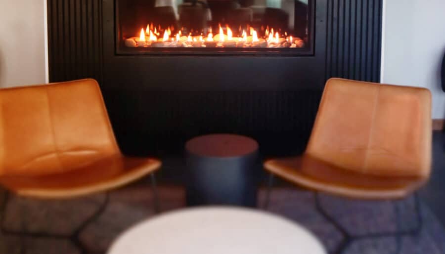 Two brown leather chairs face a modern black fireplace with a small flame. A round, white marble table is in the foreground on a textured rug. A small dark side table is between the chairs. The scene conveys a warm, minimalist setting.