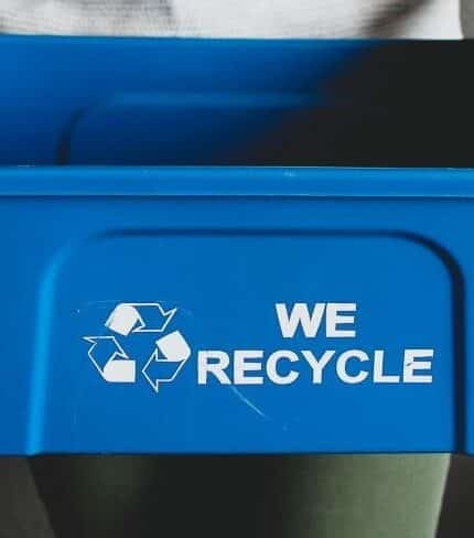 A person holds a blue recycling bin with the text "We Recycle" and a recycling symbol on the front. The person is wearing a light-colored top and has short, painted nails. The background is a dark, neutral wall.