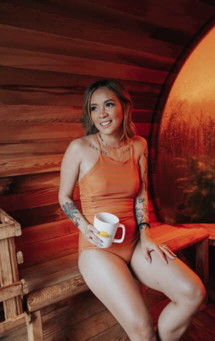 A person with tattoos sits on a wooden bench in a warm-toned room, holding a mug. They wear an orange outfit and smile, with a round, mist-covered window in the background.