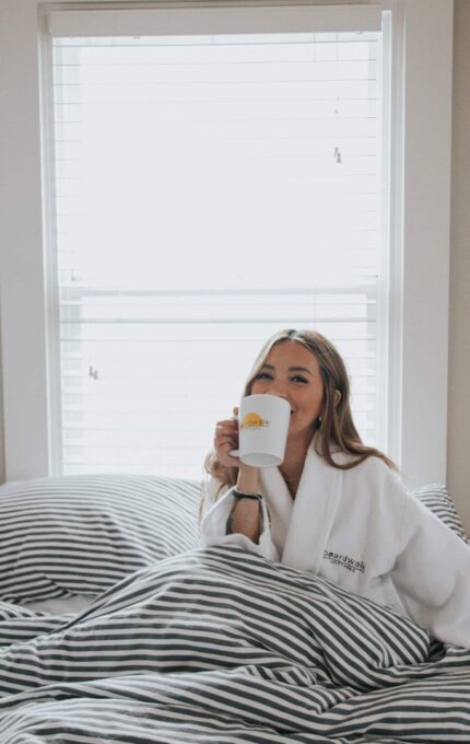 A person in a white robe sits on a striped bed, holding a mug with a sun design. They appear to be sipping from the mug, near a window with closed blinds. The setting is a bright, cozy bedroom.