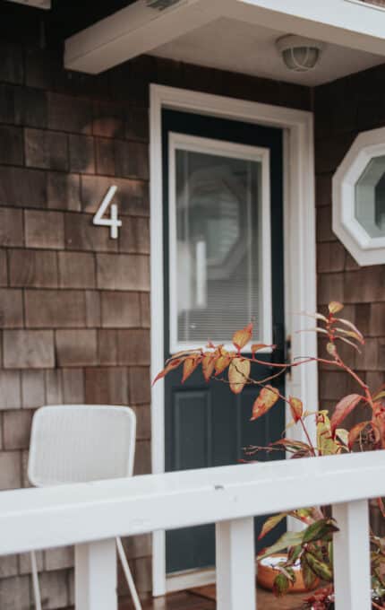 A navy blue door with a glass panel and a white frame is centered in the image. To the left, a white chair is partially visible. The exterior wall is clad in brown shingles, and the number '4' is mounted above the chair. Leaves are in the foreground.