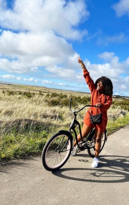 A person rides a black bicycle along a paved path through an open field with tall grass under a blue sky with scattered clouds. They wear a rust-colored outfit and are raising one arm, pointing upwards.