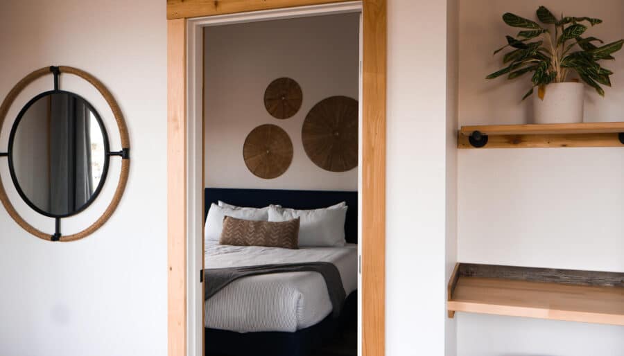 A modern bedroom is visible through a wooden-framed doorway. The room features a neatly made bed with white linens, decorative pillows, and wall-mounted woven decor. A round mirror and a small plant on a shelf are visible in the foreground.