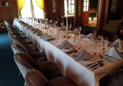 A long dining table set for a formal meal in an elegant room. The table is covered with a white tablecloth, featuring neatly folded napkins, plates, glassware, and silverware. Soft chairs surround the table. Natural light enters through large windows.