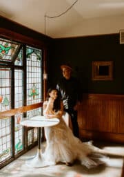 A couple stands in a warmly lit room. The woman sits at a round table wearing a white dress, while the man in a dark outfit and hat leans against the wall. Stained glass windows cast patterns on the floor.