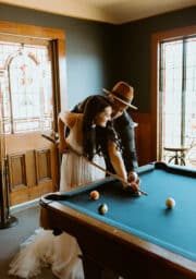 A couple in formal attire is playing pool indoors. The woman is holding a cue stick, while the man assists her with the shot. The room has stained glass windows and a wooden door in the background, and a pool table is in the foreground.