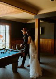 A couple stands near a pool table in a dimly lit room. The man, wearing a hat and suit, is seated on the table, while the woman in a white dress leans against him. A stained glass window and a wall-mounted TV are in the background.