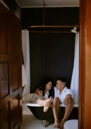 A man and woman in white robes sit together in a vintage-style bathtub with curtains, in a dimly lit bathroom. They appear relaxed and are looking at each other, surrounded by dark wood accents and a closed wooden door.