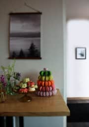 A wooden table with a stack of colorful macarons and plates of small bundt cakes with floral decorations. In the background, there is a gray-scale forest wall hanging and a framed picture on a white wall.